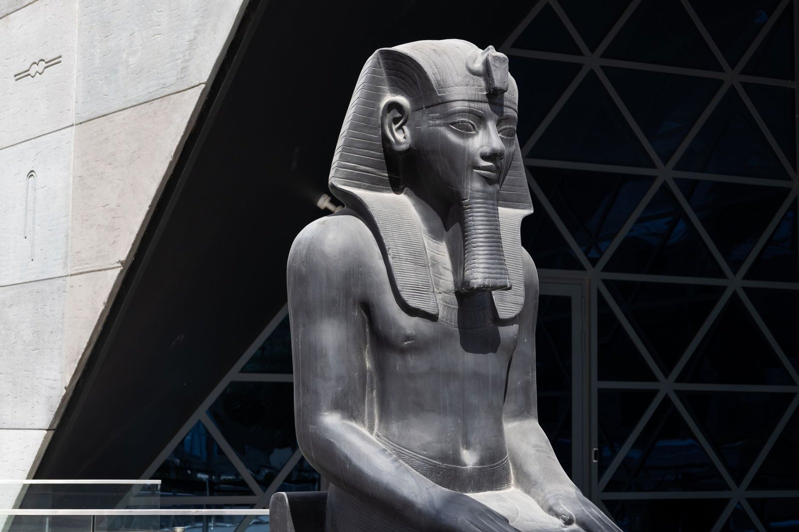 Ancient Egyptian pharaoh statue outdoors beside a modern building, with stone columns in the background