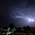 Lightning striking during a nighttime thunderstorm over a residential area, evoking Zeus’s thunderbolt