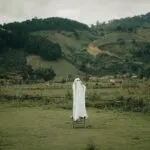 Person dressed as a ghost standing on a stool in a grassy field with mountains in the background, evoking a banshee